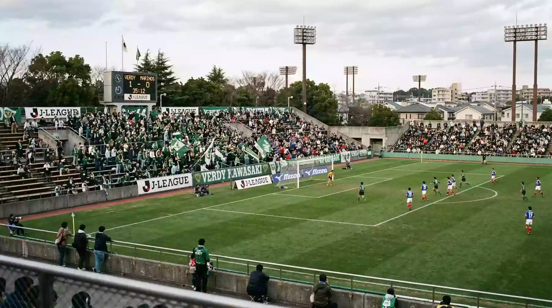 Estadio de fútbol japonés de los años noventa con jugadores en el campo durante un partido inaugural de la J-League